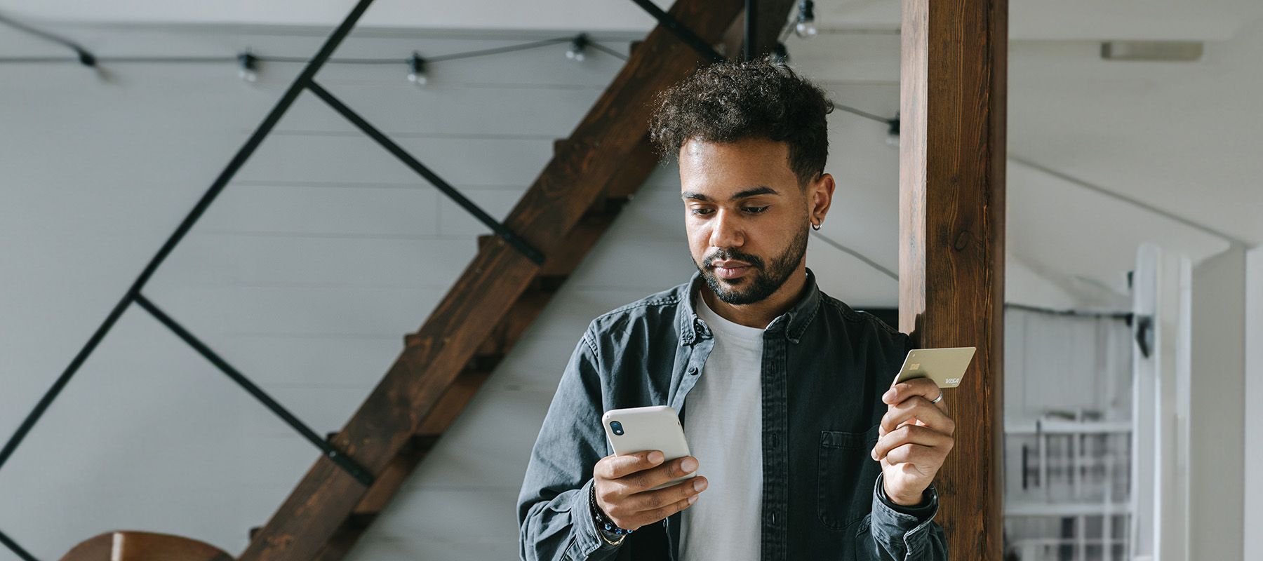 man leaning against post in loft style apartment staring at his cell phone and holding an MBNA credit card in his other hand